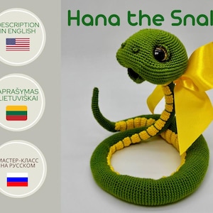 Puede incluir: Un juguete de serpiente de crochet verde con un lazo amarillo. La serpiente está enroscada en un círculo. El juguete está etiquetado como "Hana the Snake".