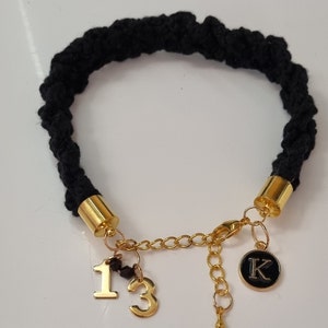 May include: Black braided bracelet with gold charms. The charms include the numbers 1 and 3, a small black bead, and a round charm with the letter K.