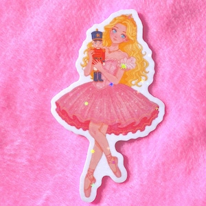May include: A die-cut sticker featuring a blonde ballerina with long wavy hair wearing a pink off-the-shoulder tutu dress and ballet slippers. The ballerina holds a small blue and red nutcracker figure. The design includes holographic star accents.
