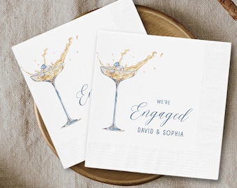 May include: Two white square napkins with a watercolor illustration of a champagne glass with a ring inside. The text "We're Engaged" is written in navy blue script, along with the names "David & Sophia".