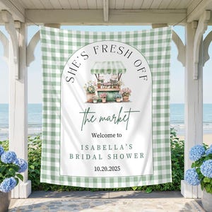 May include: A green and white gingham banner with the text "She's Fresh Off the Market" and "Welcome to Isabella's Bridal Shower 10.20.2025". The banner features a watercolor illustration of a flower stand. Blue hydrangeas in pots flank the banner.