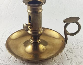 May include: Antique brass candlestick holder with a round base, a central column, and a small handle. The candlestick has a circular opening for a candle and a small knob on the side. The overall design is vintage.