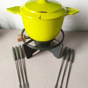 Può includere: Una pentola per fonduta in ghisa gialla con un coperchio nero e un supporto in metallo nero. La pentola è circondata da sei forchette per fonduta con manici verdi.