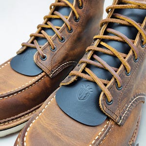 May include: A pair of brown leather moc-toe boots featuring black leather kilties with an embossed shield logo. The boots have tan leather laces, black metal eyelets, and white contrast stitching along the moccasin-style toe seam.