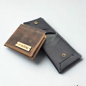 以下が含まれることがあります： さまざまな色の3つの革製財布。前面には「V-KING」と書かれた木製のタグが付いた茶色の正方形の財布があります。その背後には、スナップ留めの黒い財布があります。3番目の財布は濃い灰色です。