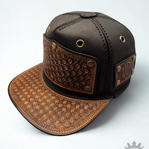 May include: A brown leather baseball cap with a decorative embossed pattern on the bill and front panel. The cap features brass eyelets and rivets, with a small logo on the side. The hat is a stylish accessory.