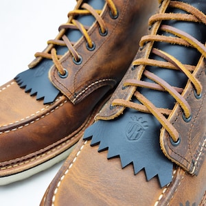 Puede incluir: Primer plano de dos botas de cuero marrón con detalles en cuero negro y costuras decorativas. Las botas tienen una puntera estilo mocasín, cordones amarillos y una franja de cuero negro. Las botas tienen un aspecto robusto y desgastado.