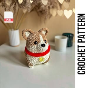 Puede incluir: Patrón de crochet PDF para un corgi amigurumi marrón y blanco con una bufanda roja y un amuleto de estrella dorada.