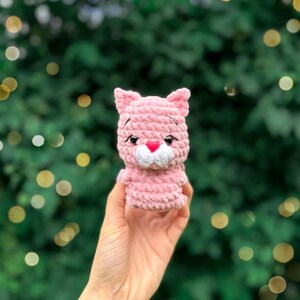 May include: A small, light pink crocheted cat figurine made from soft, plush yarn. The cat features pointed ears, embroidered black eyebrows, black plastic safety eyes, and a small, bright pink heart-shaped nose above a white muzzle.
