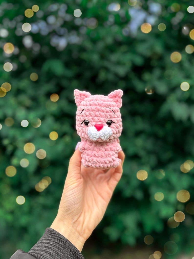 May include: A small, light pink crocheted cat figurine made from soft, plush yarn. The cat features pointed ears, embroidered black eyebrows, black plastic safety eyes, and a small, bright pink heart-shaped nose above a white muzzle.