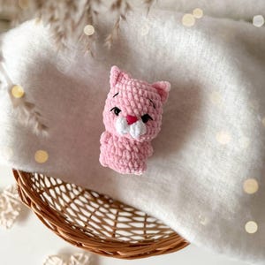 May include: A small, light pink crocheted cat figurine made from soft, plush yarn. The cat features embroidered black eyes, a small pink heart-shaped nose, and a white muzzle area. It has pointed ears and a rounded body, crafted with a textured stitch.