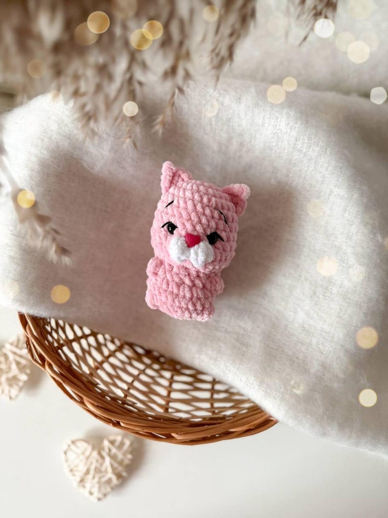 May include: A small, light pink crocheted cat figurine made from soft, plush yarn. The cat features embroidered black eyes, a small pink heart-shaped nose, and a white muzzle area. It has pointed ears and a rounded body, crafted with a textured stitch.