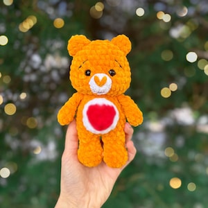 May include: An orange plush bear with a white belly and a red heart. The bear has small black eyes and a white heart-shaped nose. The bear is being held in a hand against a blurred green background.