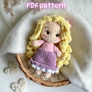May include: A crochet doll with long, curly yellow yarn hair, a light pink long-sleeved top, and a purple skirt with a white ruffled hem. The doll has embroidered facial features and is labeled with the text PDF pattern at the top.