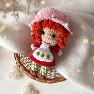 May include: A crocheted doll with bright red curly hair, wearing a pink hat with small white pom-poms, a red dress, and a white apron featuring green strawberry embroidery. The doll has green and white checkered leggings and brown shoes.