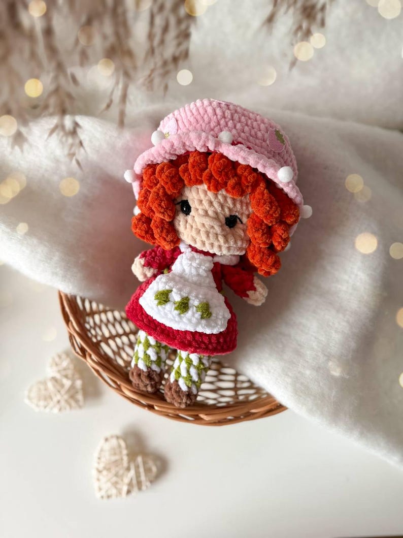 May include: A crocheted doll with bright red curly hair, wearing a pink hat with small white pom-poms, a red dress, and a white apron featuring green strawberry embroidery. The doll has green and white checkered leggings and brown shoes.