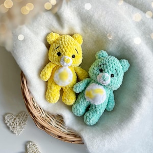 May include: Two handmade crochet teddy bears in a wicker basket. One bear is yellow, the other is teal. Both have white and yellow accents on their bellies. The bears are sitting on a white blanket.