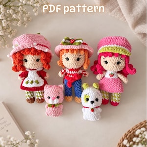 May include: Three crocheted dolls and two small crocheted animals. The dolls wear hats and colorful outfits with strawberry accents. The animals are a pink cat and a white dog. The text PDF pattern is visible at the top of the image.