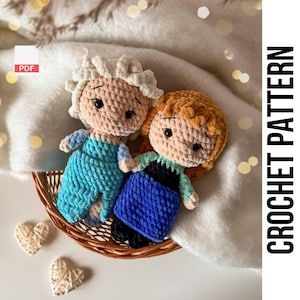 May include: Two crocheted dolls, one with a blue dress and blonde hair, the other with a blue and green dress and brown hair. The dolls are sitting in a wicker basket.