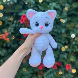 Puede incluir: Mu&ntilde;eca de gato de ganchillo blanco hecha a mano con detalles rosas en las orejas, la nariz y las patas. El gato tiene grandes ojos azules y un peque&ntilde;o lazo morado. La mu&ntilde;eca se sostiene contra un fondo verde y rojo borroso.