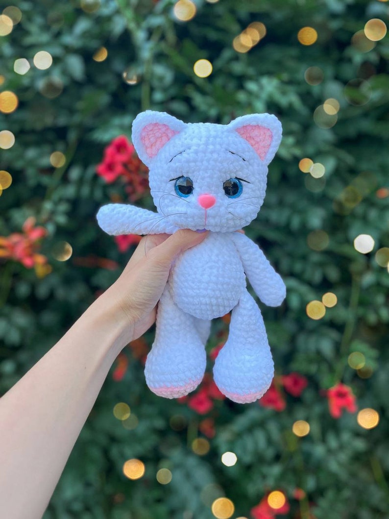 Puede incluir: Mu&ntilde;eca de gato de ganchillo blanco hecha a mano con detalles rosas en las orejas, la nariz y las patas. El gato tiene grandes ojos azules y un peque&ntilde;o lazo morado. La mu&ntilde;eca se sostiene contra un fondo verde y rojo borroso.