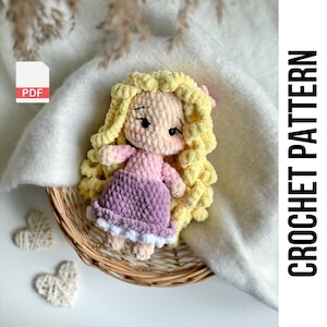 May include: Crochet pattern PDF for a doll with long blonde yarn hair, wearing a purple dress with a white trim, and a pink shirt. The doll is sitting in a wicker basket.