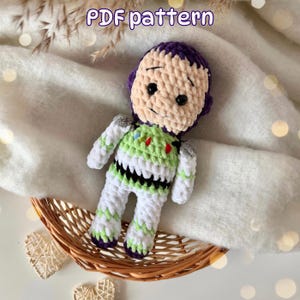 May include: A crochet doll featuring a character with a purple hood, beige face, and black safety eyes. The body is white with lime green and black accents, including small red and blue details on the chest. The text PDF pattern is visible.