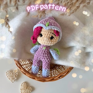 Puede incluir: Una muñeca de ganchillo hecha a mano con un atuendo rosa y morado, detalles azules y alas verdes. La muñeca tiene la piel clara, ojos negros y pelo rosa. El texto "PDF pattern" está escrito en rosa.