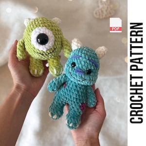 Könnte beinhalten: Zwei handgefertigte Häkeltier-Plüschtiere, eines grün mit einem großen schwarzen Auge und das andere blau mit lila Markierungen. Die Spielzeuge werden in Händen gehalten. Der Text "CROCHET PATTERN" ist vertikal in Schwarz gedruckt.