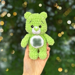 May include: A handmade, green crochet bear with black eyes and a white heart-shaped patch on its chest. The bear is held in a hand against a blurred green and gold background. The bear's fur is a soft, textured yarn.