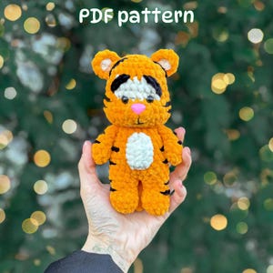 May include: A crochet tiger plush toy featuring bright orange yarn, black stripes, a white belly patch, and a white face patch with black embroidered eyebrows and a pink plastic nose. The text PDF pattern is displayed at the top of the image.