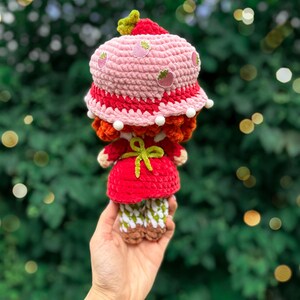 May include: A crocheted doll wearing a pink strawberry-patterned bucket hat with white pom-pom trim, a red dress with a bright green tied apron, and white and green checkered leggings with brown shoes, featuring textured yarn construction throughout.