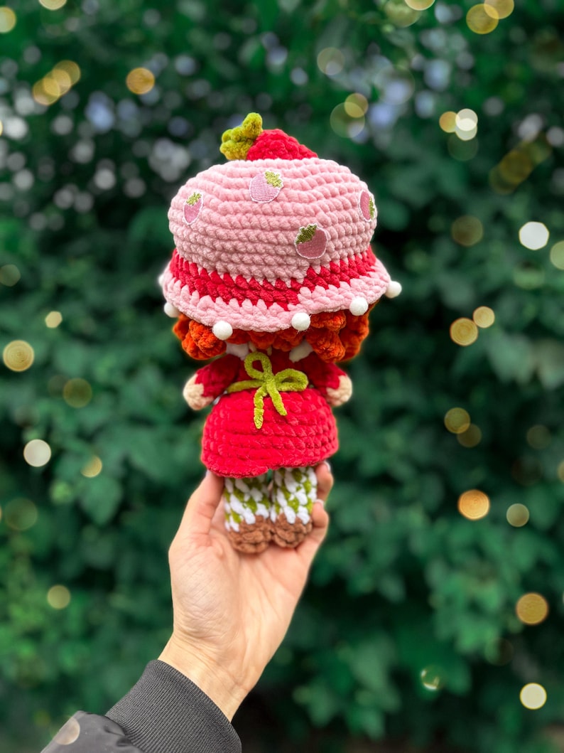 May include: A crocheted doll wearing a pink strawberry-patterned bucket hat with white pom-pom trim, a red dress with a bright green tied apron, and white and green checkered leggings with brown shoes, featuring textured yarn construction throughout.