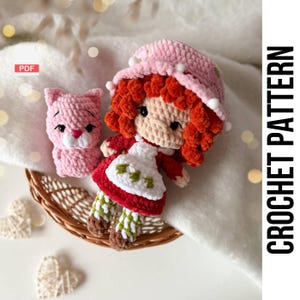 May include: A crochet doll and a pink cat are displayed in a woven basket. The doll has red hair, a pink hat, and a red and white dress. The cat is pink. The words "CROCHET PATTERN" are printed vertically in black.