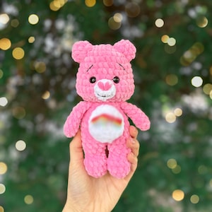 May include: A handmade pink teddy bear with a white belly featuring a rainbow design. The bear has black eyes, a pink heart-shaped nose, and a white embroidered smile. The plush toy is held in a hand against a blurred green background.