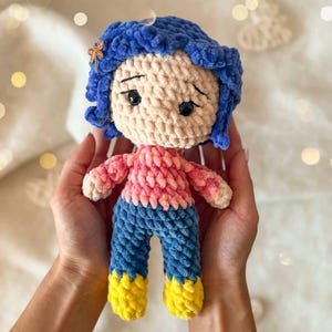 May include: A handmade crochet doll featuring bright blue yarn hair with a small dragonfly hair clip, a light beige face with black embroidered eyes, a pink textured sweater, blue pants, and yellow boots, crafted from soft plush yarn material.
