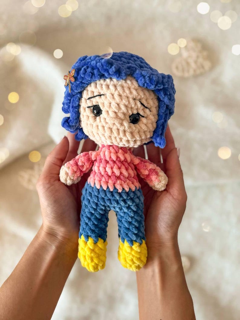 May include: A handmade crochet doll featuring bright blue yarn hair with a small dragonfly hair clip, a light beige face with black embroidered eyes, a pink textured sweater, blue pants, and yellow boots, crafted from soft plush yarn material.