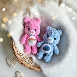 May include: Two handmade crochet teddy bears, one pink and one blue, rest in a woven basket on a white surface. The bears have embroidered features and a cloud-like design on their bellies. Soft, fluffy texture.