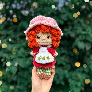 May include: A handmade crochet doll featuring bright orange yarn hair, a light pink bucket hat with small white pom-poms and strawberry accents, a red dress, a white apron with green leaf patterns, and white and green patterned leggings.