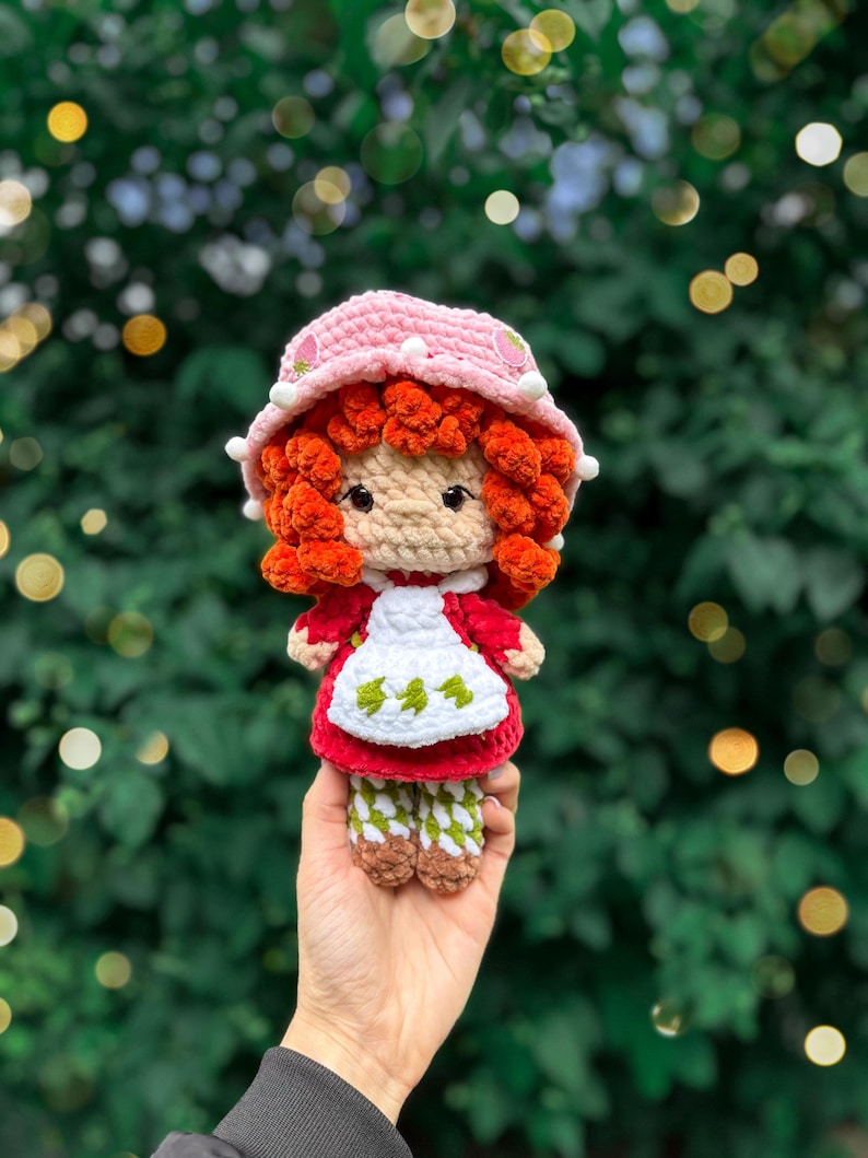May include: A handmade crochet doll featuring bright orange yarn hair, a light pink bucket hat with small white pom-poms and strawberry accents, a red dress, a white apron with green leaf patterns, and white and green patterned leggings.
