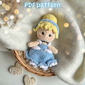 May include: A crochet doll featuring a light blue dress with white trim, yellow hair styled in an updo, and a blue headband. The doll has a beige face with embroidered eyes and pink cheeks. The text "PDF pattern" is visible at the top.