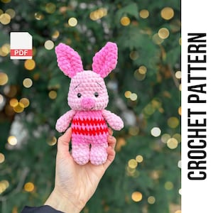 Könnte beinhalten: Ein rosa gehäkeltes Plüschtier Piglet mit rot-rosa gestreiften Kleidung. Das Spielzeug wird in einer Hand vor einem verschwommenen grünen Hintergrund gehalten.