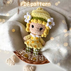 May include: A crochet doll with yellow hair, a yellow dress with orange dots, and white flower accents on the head. The doll has black eyes and wears green and white patterned leggings with small beige bows. The text PDF pattern is visible.