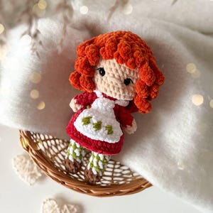 May include: A handmade crochet doll featuring vibrant orange curly hair, a beige face with black eyes, and a red dress with a white apron. The apron is decorated with small green leaf patterns. The doll wears green and white checkered leggings and brown shoes.