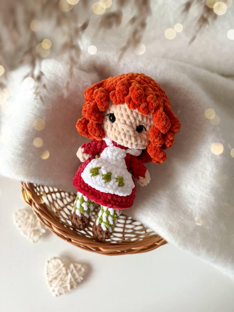 May include: A handmade crochet doll featuring vibrant orange curly hair, a beige face with black eyes, and a red dress with a white apron. The apron is decorated with small green leaf patterns. The doll wears green and white checkered leggings and brown shoes.