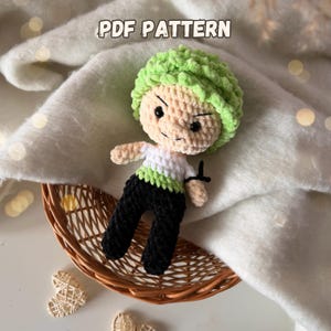 Könnte beinhalten: Gehäkelte Puppe einer Figur mit grünen Haaren, schwarzer Hose und einem weiß-grünen Hemd. Die Puppe befindet sich in einem Weidenkorb. Der Text "PDF PATTERN" befindet sich oben im Bild.