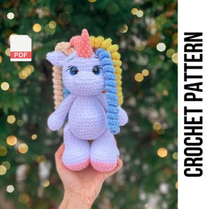Może przedstawiać: Wzór na szydełku na białego jednorożca amigurumi z różowym rogiem i tęczową grzywą z włóczki. Wzór dostępny jest jako plik PDF do pobrania.