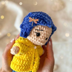 May include: A handmade crochet doll featuring a plush blue bob hairstyle adorned with a small gold dragonfly hair clip. The doll wears a bright yellow textured coat over blue pants and has a light beige face with black button eyes and stitched brows.