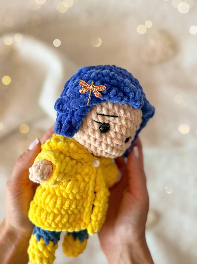 May include: A handmade crochet doll featuring a plush blue bob hairstyle adorned with a small gold dragonfly hair clip. The doll wears a bright yellow textured coat over blue pants and has a light beige face with black button eyes and stitched brows.