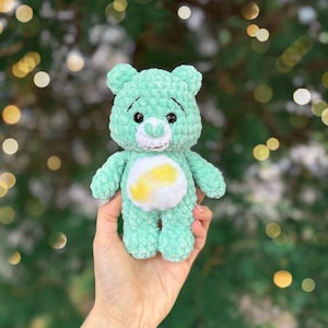 May include: A mint green crocheted bear plushie with a white and yellow belly. The bear has black eyes and a white muzzle. The plush toy is held in a hand against a blurred green and gold background.